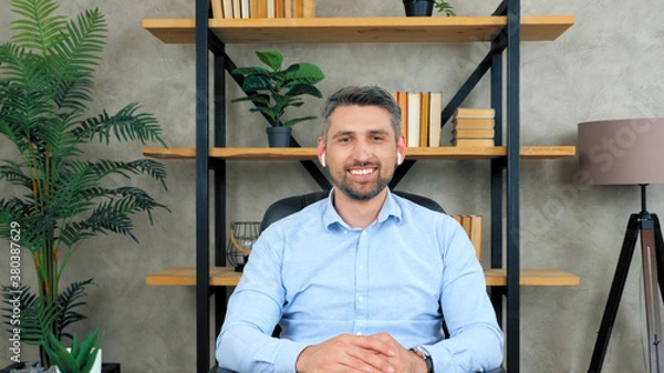 Fototapeta Smiling positive happy businessman sitting on chair at table in home office wearing wireless earphones looks into camera listening to interlocutor on video call chat conference, web cam concept