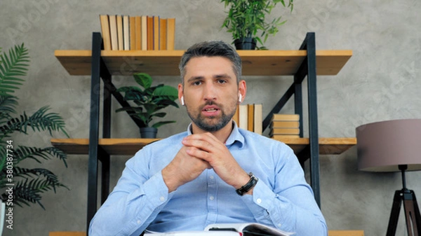 Fototapeta Confident serious man remote teacher online tutor in wireless earphones looks camera teaches communicates students video call chat conference web cam laptop computer view, distance education learning