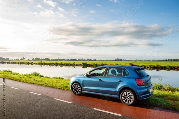 Fototapeta A blue parked car on the side of the road near the ringvaart canal in the North-Holland village of Abbenes on a cloudy day in the Netherlands.