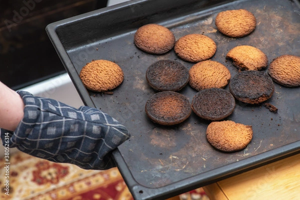 Obraz Burnt cookies. A hand in a oven glove, potholder picks up a black baking tray with burnt cookies