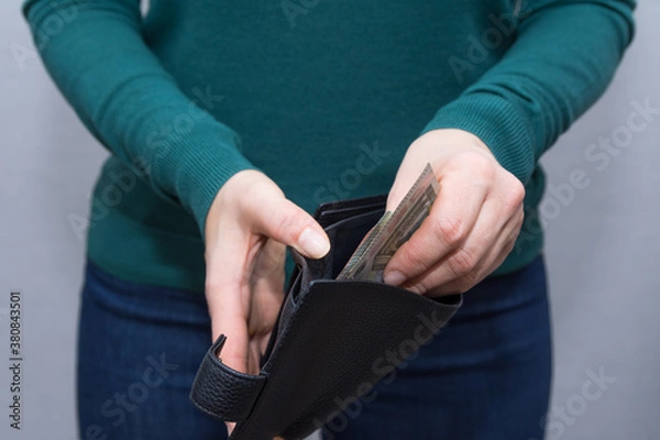 Fototapeta Woman in green turtleneck stands over grey background. She holds good expensive leather wallet and gets some euro money out oit. Modern wallet made of black genuine leather with some banknotes inside.