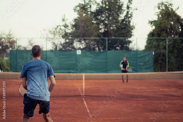 Fototapeta Man in blue shirt and blue pants seen from behind playing tennis, wide open clay orange playing field. Anticipating the serve from the other side, outline of the opponent hitting a ball