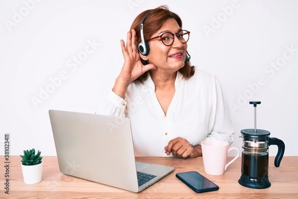 Fototapeta Middle age brunette hispanic woman working at the office wearing operator headset smiling with hand over ear listening and hearing to rumor or gossip. deafness concept.
