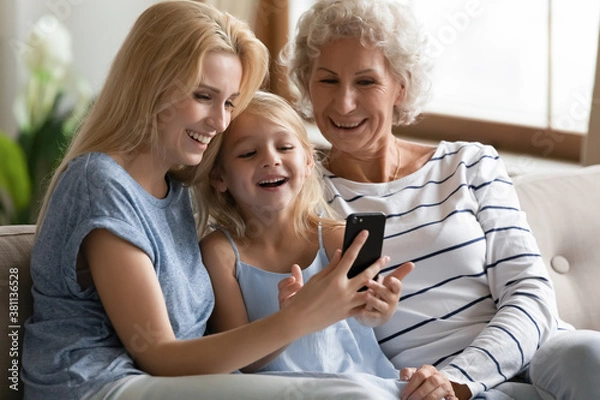 Fototapeta Cute thing. Happy laughing little kid girl relaxing on couch on weekend with senior grandmother and adult mom reading internet blog on cell, playing online game, sharing photos, enjoying video by wifi