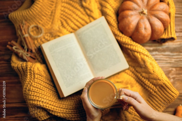 Fototapeta Hot cocoa in hands, ripe pumpkin, warm sweater and open book on wooden background. Vintage book with romantic stories and fairy tales. Top view of autumn composition with book and knitted sweater.