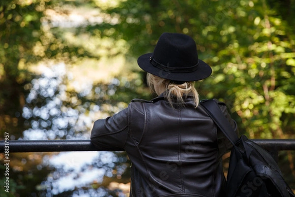 Fototapeta young Caucasian woman in a leather jacket, hat and with a backpack stands on the bridge and looks at the river. selective focus. High quality photo