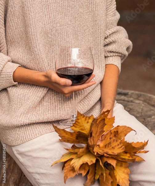 Fototapeta A women's hands holding a glass of red wine and an autumn bouquet of yellow fall leaves. Beige trendy total look with warm sweater and trousers. Trendy autumn concept in pastel colors. Autumn mood.