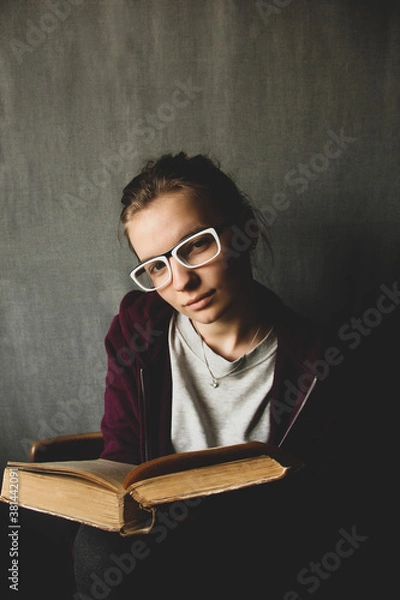 Obraz Girl in glasses with a book on a gray background. The girl is holding an old book.
