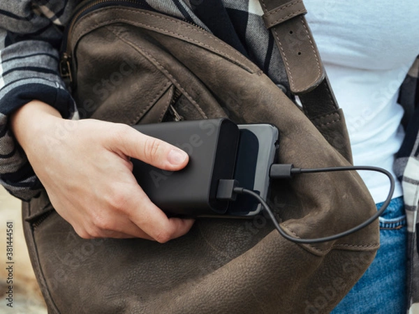 Fototapeta The girl uses a smartphone connected to the power Bank. Black smartphone and powerBank in women's hands close-up. Using gadgets in the modern world. The concept of portable chargers, modern tourism