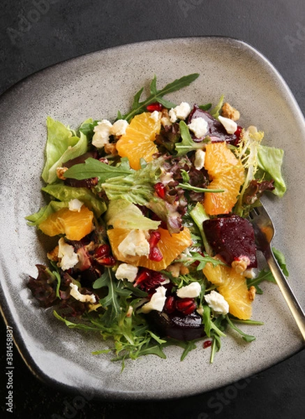 Fototapeta Salad with arugula, orange, beet, cheese, pomegranate seeds and nuts on a gray plate on a black background. Background image, copy space
