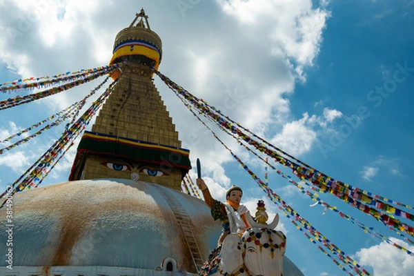 Fototapeta The upper part of the Boudhanath stupa in Kathmandu, Nepal. The image of Buddhas eyes on a gold surface of the stupa. Colorful prayer flags nearby. Buddha statue on a white elephant.