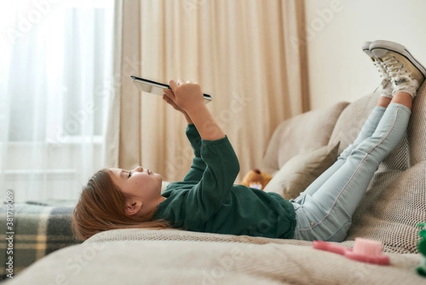 Obraz A small cute girl playing games on a tablet lying on a sofa with her feet up and gumshoes on