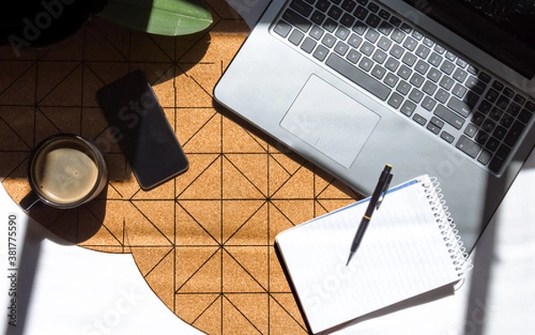Obraz Vista cenital de un escritorio moderno con una plancha de corcho de figuras geométricas bajo la luz del sol de la mañana. Oficina en el hogar con laptop, teléfono móvil, anotador y una taza de café.