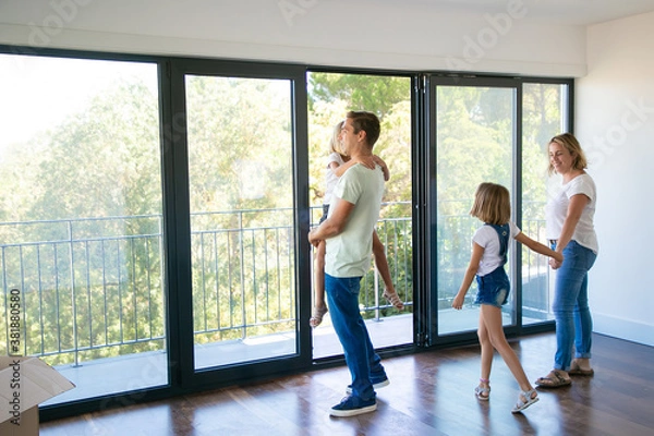 Obraz Happy father with daughter standing near open balcony and smiling. Blonde mother holding girl hand and talking with her. Enjoying family watching new house or flat. Relocation and mortgage concept