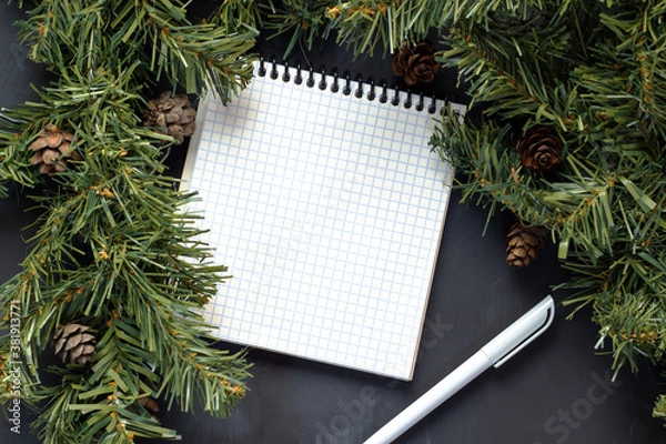 Obraz A paper notebook with a white pen lies on a black background surrounded by spruce branches with cones.