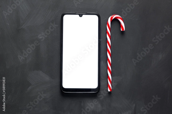 Obraz A smartphone with a white screen lies on a dark background next to a red candy cane.