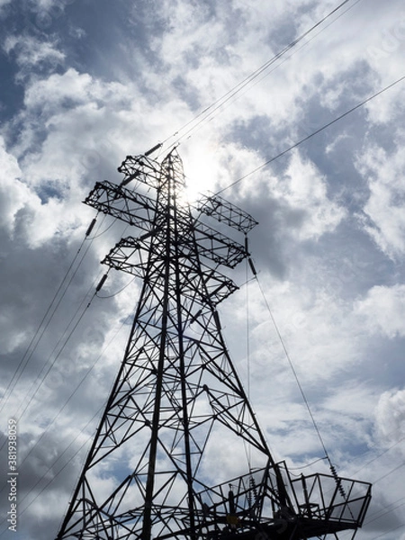 Fototapeta An electricity pylon viewed from its base looking upwards into a cloudy sky with the sun peeping through the clouds
