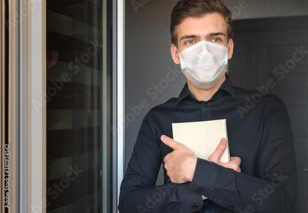 Obraz Portrait of a young man in a protective medical mask, at the open door, with a book in his hands. The concept of quarantine, online training, and the  second wave of coronavirus.