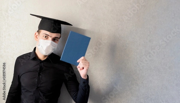 Obraz Portrait of a young student in a graduation cap and protective mask with a diploma in his hand, on a gray background, banner. The concept of distance education in the context of the coronavirus pandem