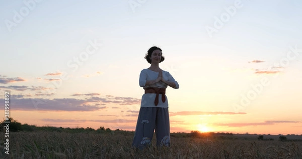 Fototapeta Female practicing qigong in summer fields with beautiful sunset on background	