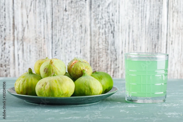 Fototapeta Green figs in a plate with drink side view on plaster and wooden background