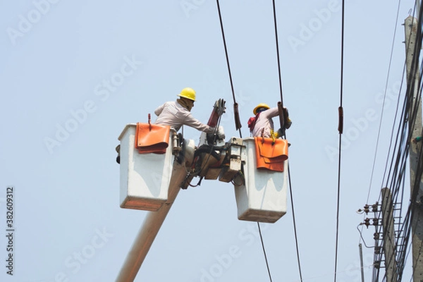Fototapeta Electricians work together on the electric cable car and electric pole. To maintain the high and low voltage distribution system. They wear a helmet with PPE protection and copy space for your text.