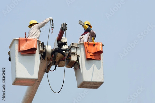 Fototapeta Electricians work together on the electric cable car and electric pole. To maintain the high and low voltage distribution system. They wear a helmet with PPE protection and copy space for your text.