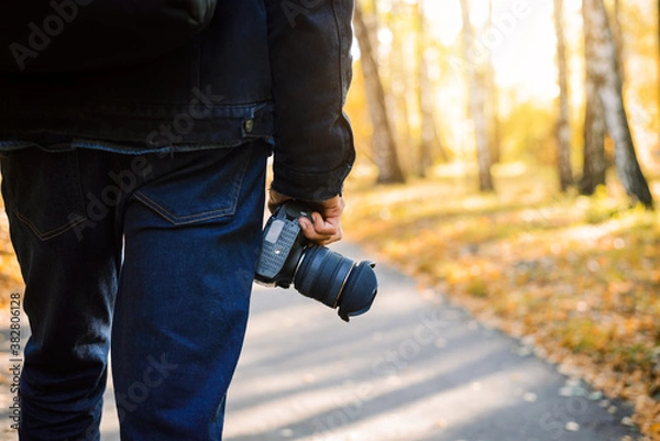 Fototapeta Man photographer from his back, walking in the park with camera in his hands. Autumn view on the background.