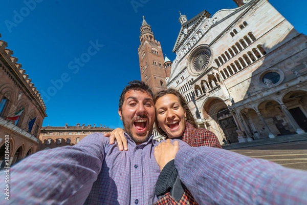 Fototapeta happy tourist couple visiting the city of Cremona taking selfie in the middle of main square of Cremona with Cathedral, baptistery and Torrazzo bell tower . 