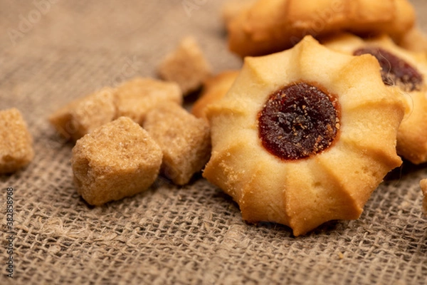Fototapeta Homemade pastry cookies with jam and pieces of brown cane sugar on a background of homespun fabric with a rough texture, close-up, selective focus.