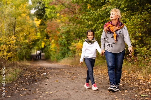 Fototapeta Happy senior lady and a little toddler girl, grandmother and granddaughter, enjoying a walk in the park. Child and grandparent. autumns day. Grandmother and little girl happy together in the garden