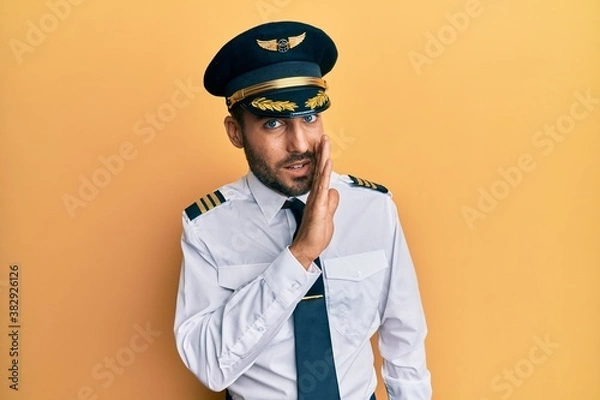 Fototapeta Handsome hispanic man wearing airplane pilot uniform hand on mouth telling secret rumor, whispering malicious talk conversation