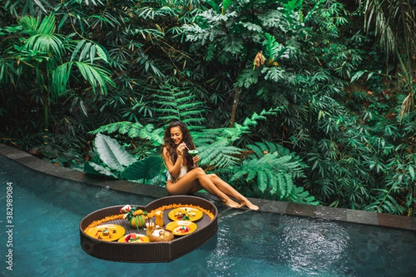 Fototapeta Girl relaxing and eating floating breakfast in jungle pool on luxury villa in Bali. Valentines day or honeymoon surprise. Tropical travel lifestyle. Black rattan tray in heart shape.