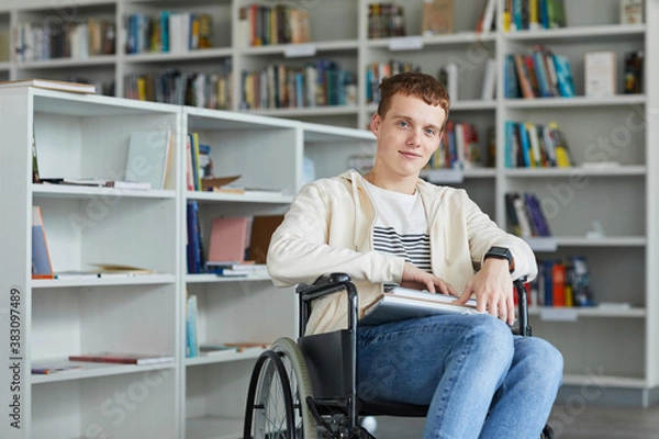 Fototapeta Portrait of smiling young man using wheelchair in school library and looking at camera, copy space
