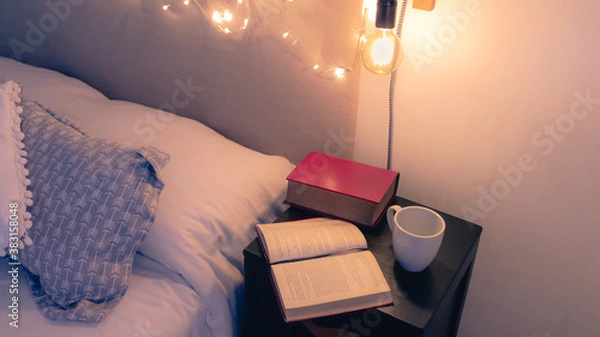 Obraz Leer un buen libro antes de dormir. Plano levemente elevado de mesa de noche junto a una cama con libros antiguos y una taza de café en un cálida confortable e iluminada habitación. 