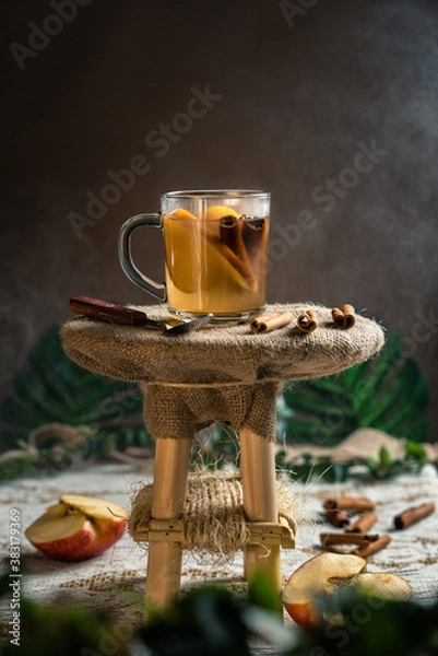 Fototapeta Cinnamon and apple tea on a rustic wooden stand with apples and cinnamon sticks in the table, steam in the dark background with copy space.