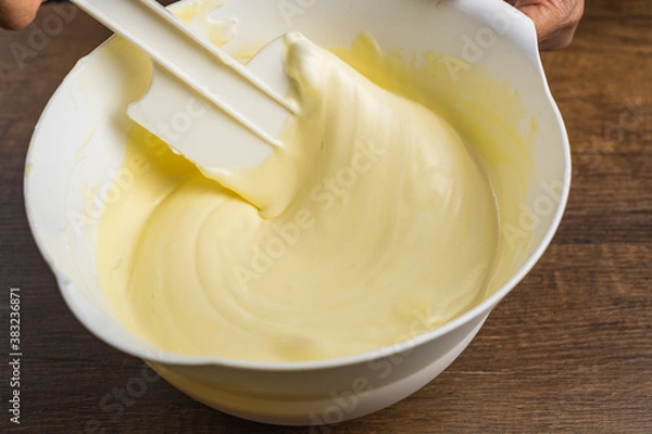 Obraz Folding smooth light yellow cake mixture with a spatula in a white bowl on top of wood table background.