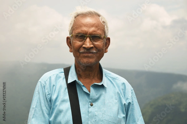 Obraz An old man, wearing spectacles and blue shirt, standing alone in nature a blurry mountain background. The elder person with a side bag on his shoulder, looking towards the camera. Copy Space For Text.