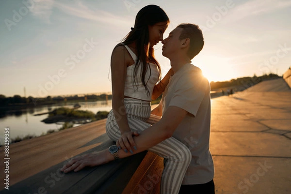 Fototapeta A young couple is sitting on a bench at sunset. The lady sits on the guy's lap and looks so contented and happy.
