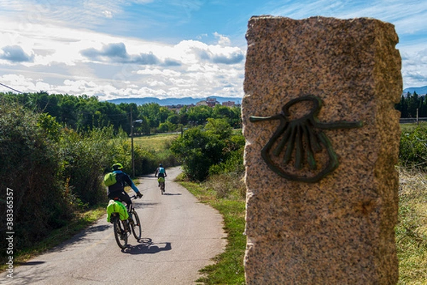 Fototapeta Pilgrims cyclists on the route of the Camino de Santiago (Way of Saint James) passing next to the scallop shell that marks the way