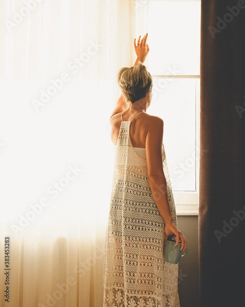Fototapeta Adult woman looking out of the window. View from the back with cup in her hand.
