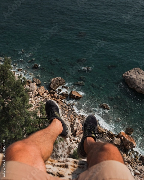 Fototapeta Legs of man are hanging from edge of cliff over the sea and rocks.
