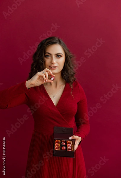 Obraz Portrait of woman in red dress posing with little box of bonbons and looking at camera. Beautiful female holding one round chocolate candy. Conept of Christmas and New Year holidays.