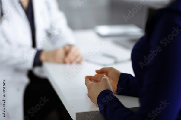 Fototapeta Unknown woman-doctor is talking to her patient about her diagnosis, while sitting together at the desk in the cabinet in a clinic. Physician at working place, close-up. Perfect medical service in a