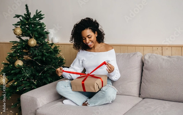Obraz Smiling african american young woman opens box with christmas gift. Christmas lifestyle and shopping concept. Black friday sale.