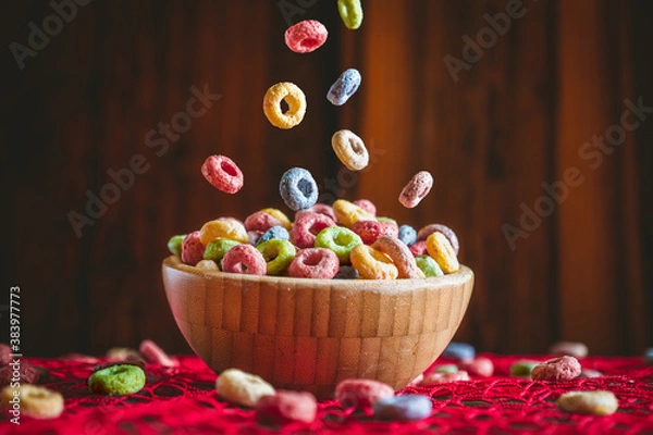 Fototapeta Colorful round fruit cereals falling into a wooden bowl on a red table. Preparing breakfast. 