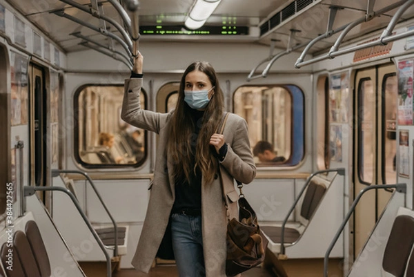Obraz A woman in a face mask to avoid the spread of coronavirus is holding onto the handrail and posing in the center of the subway car. Girl in a surgical mask against COVID-19 is standing on a metro train