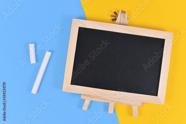 Fototapeta Empty wooden black chalkboard next to pieces of white chalk on yellow and blue background