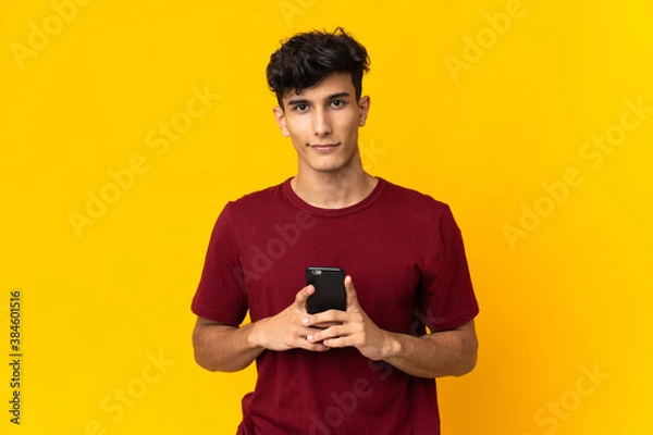 Fototapeta Young Argentinian man isolated on yellow background looking at the camera and smiling while using the mobile