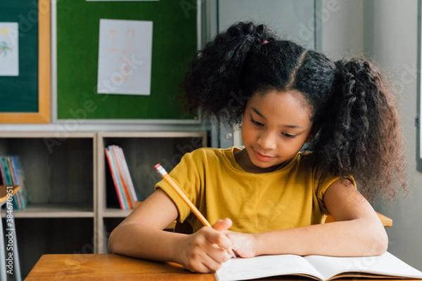 Obraz African American school girl sitting in school writing in note book with pencil, studying, education, learning. Female student at desk in classroom in exam.